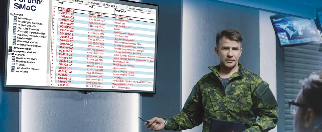 A Soldier in a modern operation centre standing next to a screen displaying a table of SMaC. Additional screens and soldiers working on computers are visible in the background.