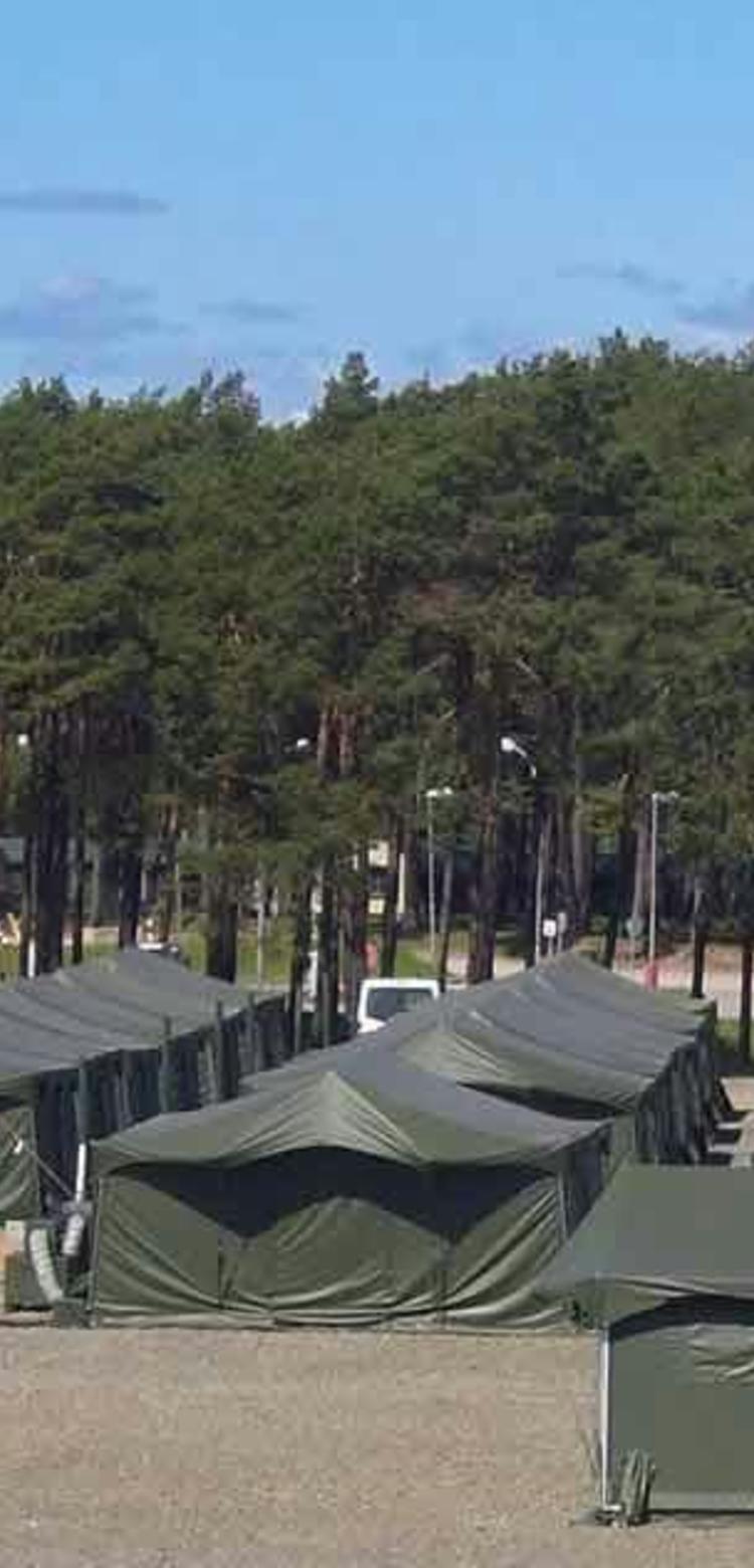 Military tent and container based infrastructure on sandy ground in front of a forest 